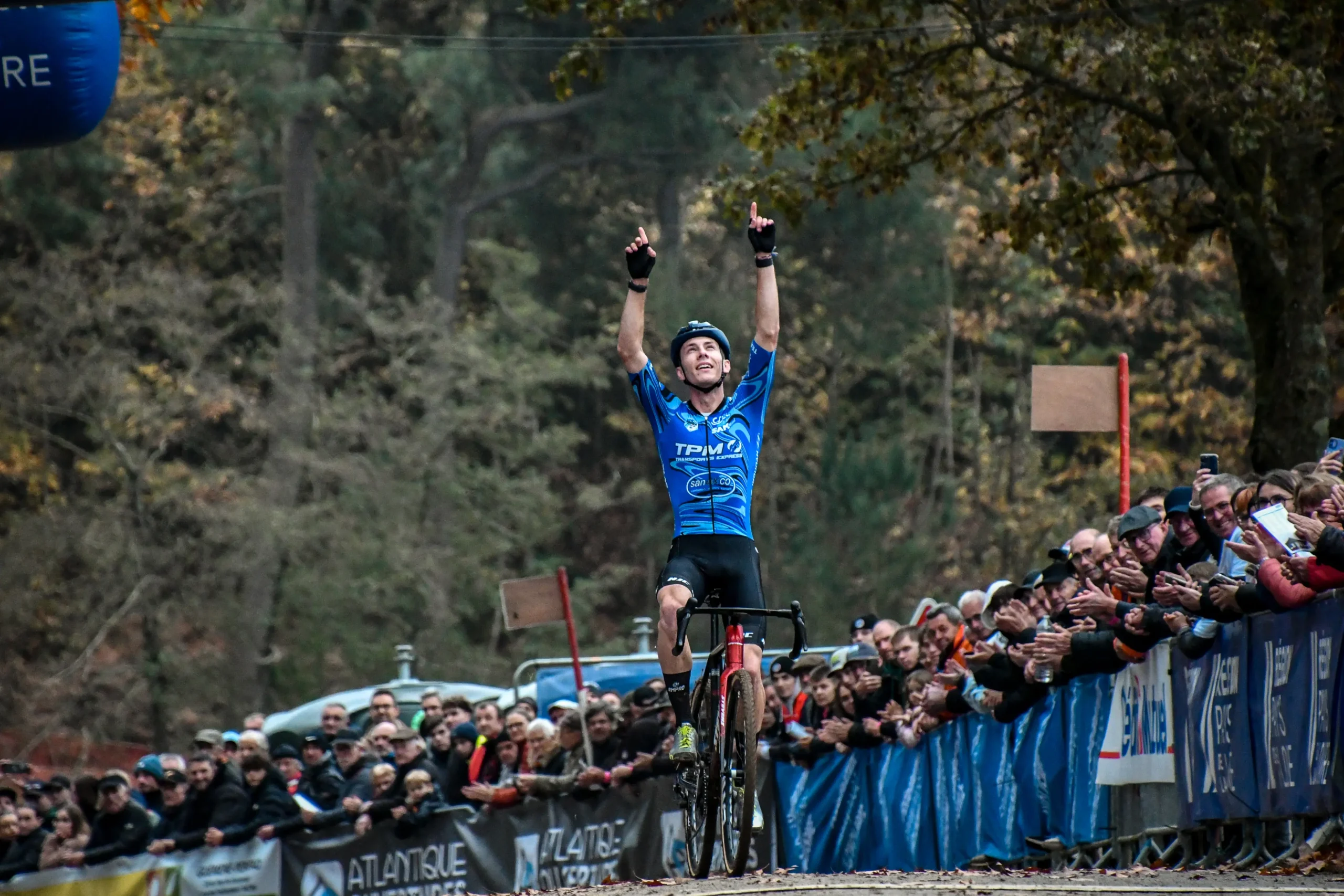 Cycliste en tenue personnalisée bleue levant les bras en signe de victoire devant un public enthousiaste.