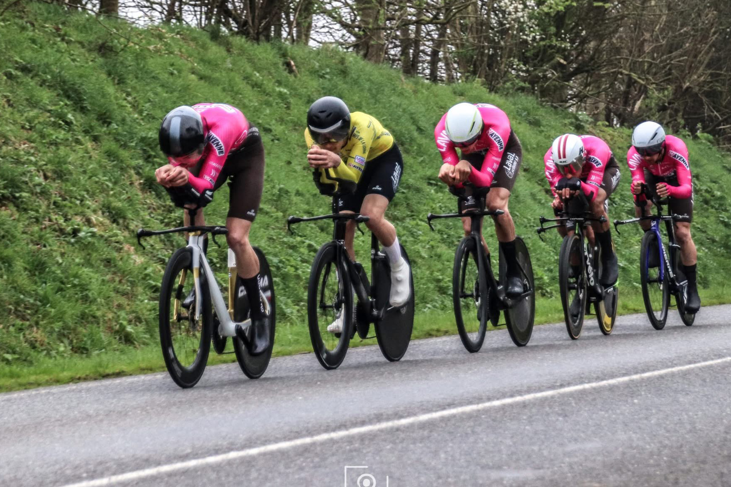 Quintette de cyclistes en tenue rose et jaune manœuvrant sur route, concentrés en position aérodynamique.